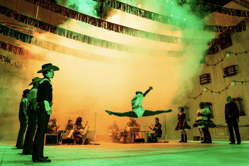 Production photo, actors stand on the side, dancer in mid air in center